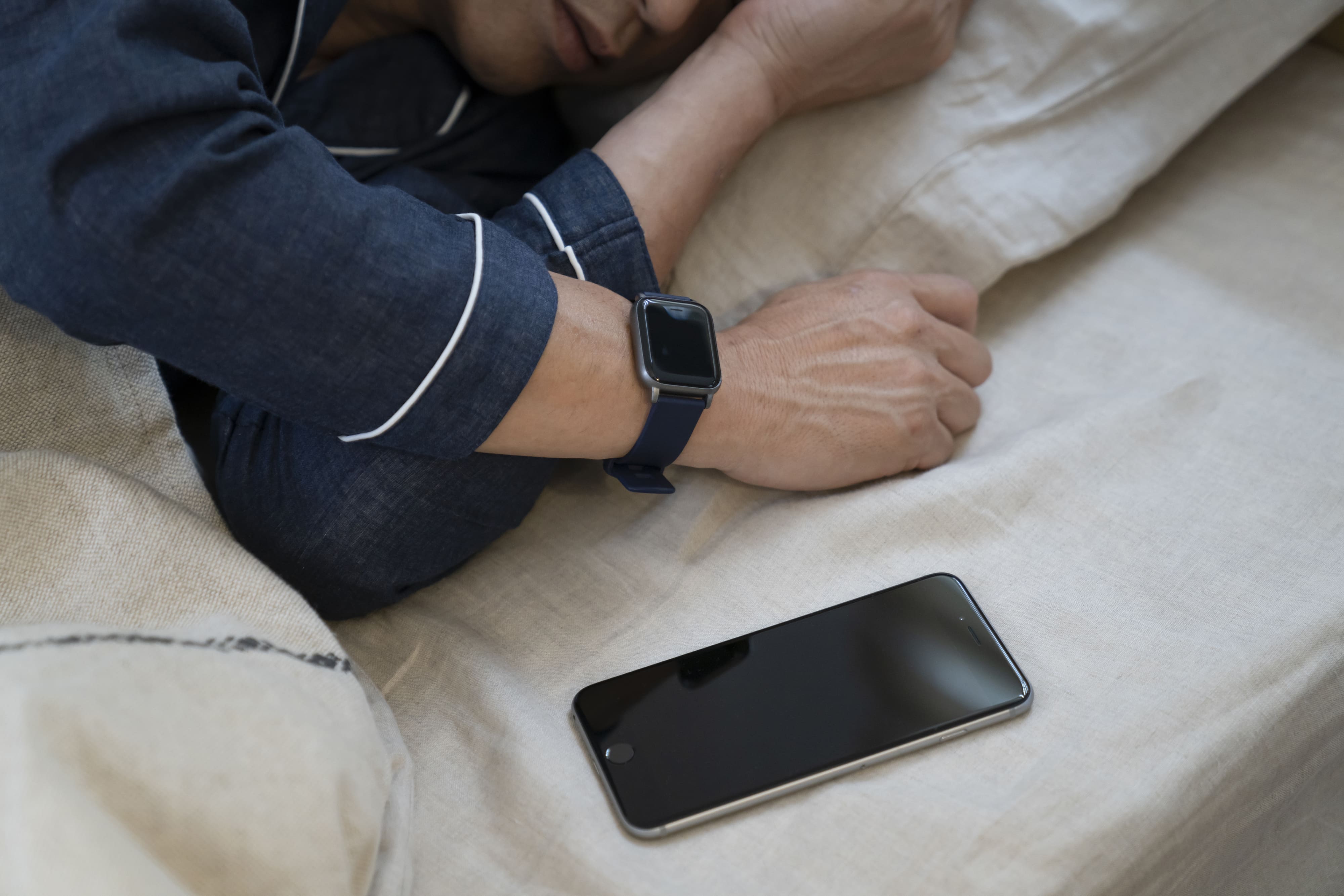 手首にスマートウォッチを身につけてベッドに横たわる人と横に置かれたスマートフォン